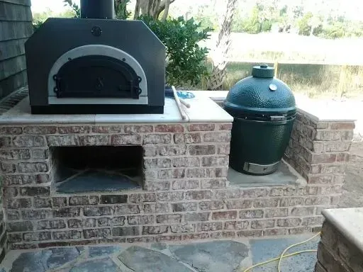 A pizza oven is built into a brick wall next to a green egg grill.