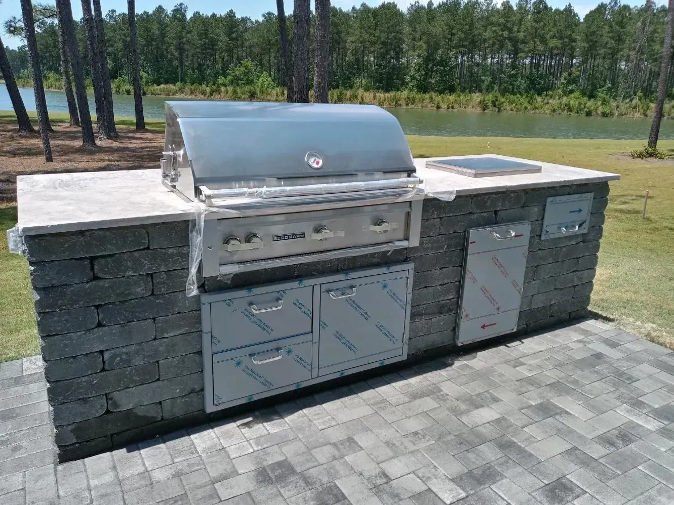 A stainless steel grill is sitting on a brick patio next to a lake.