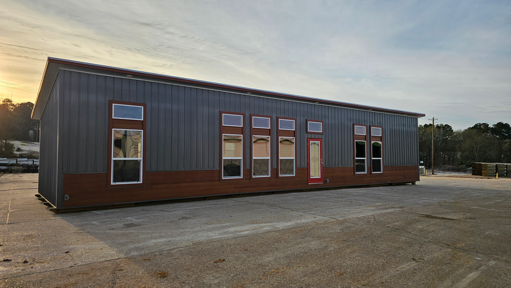 Gray and brown modular home with multiple windows and a red door, outdoors.