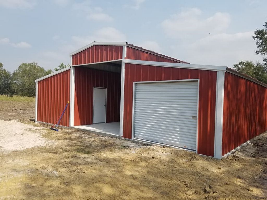Red metal barn with open carport and garage door, set in a dirt lot under a blue sky.
