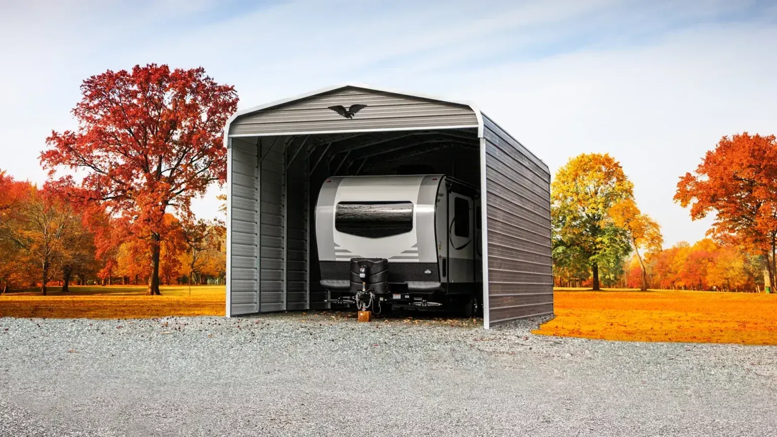 Gray RV inside a gray metal shelter, set on gravel driveway, autumn trees.