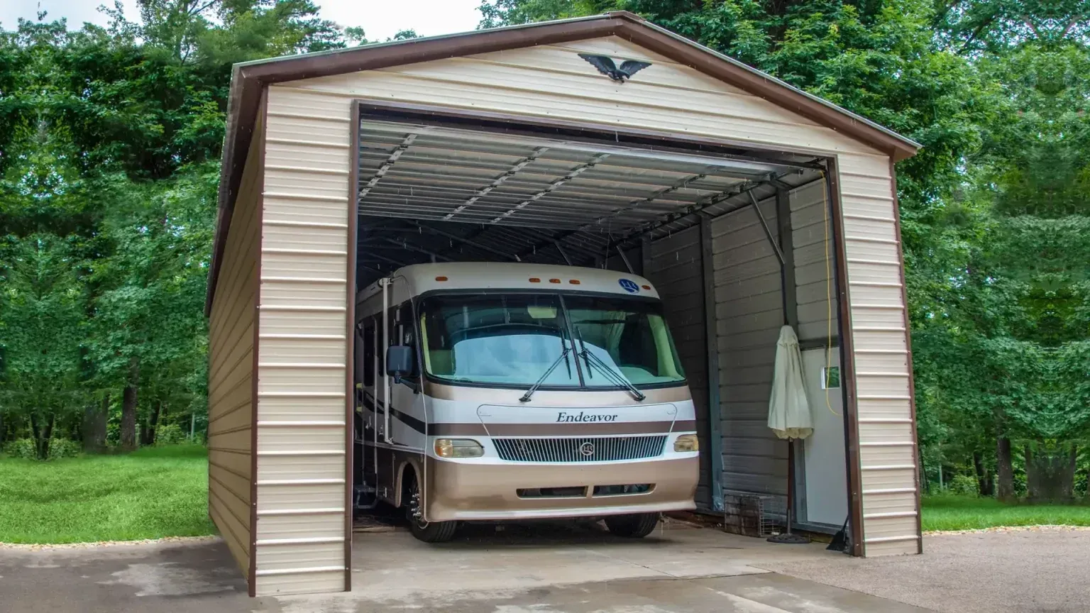 RV parked in a tan metal garage with brown trim, set against a backdrop of trees.