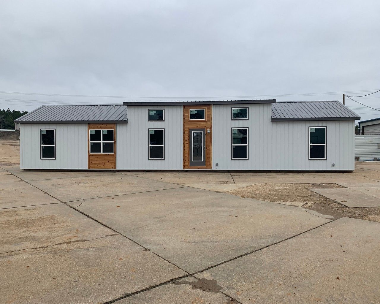 A long, light-gray manufactured home with wood accents and a dark metal roof, sitting on a concrete pad.