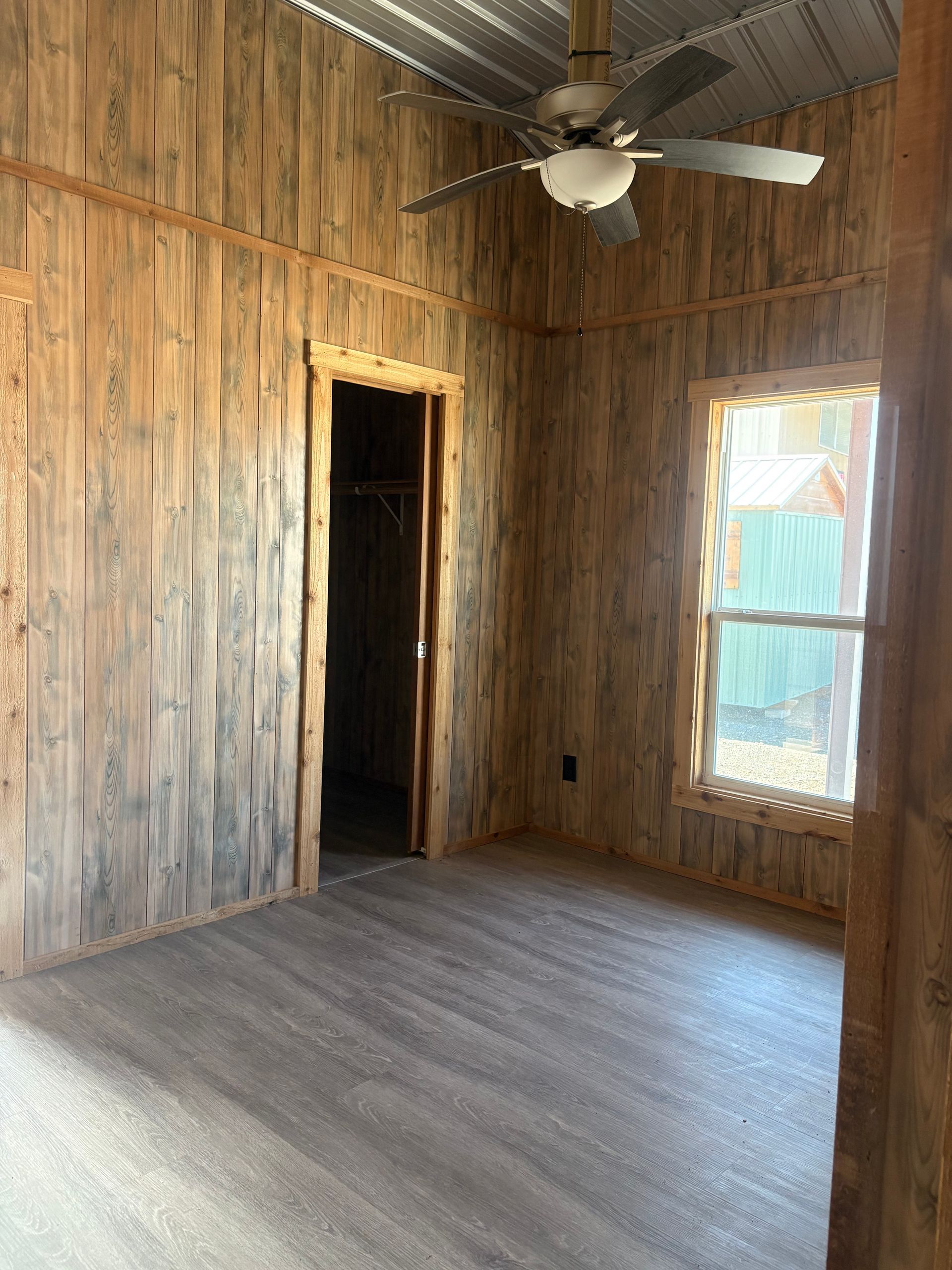 A rustic bedroom with wooden walls and flooring, a closet, a window, and a ceiling fan.