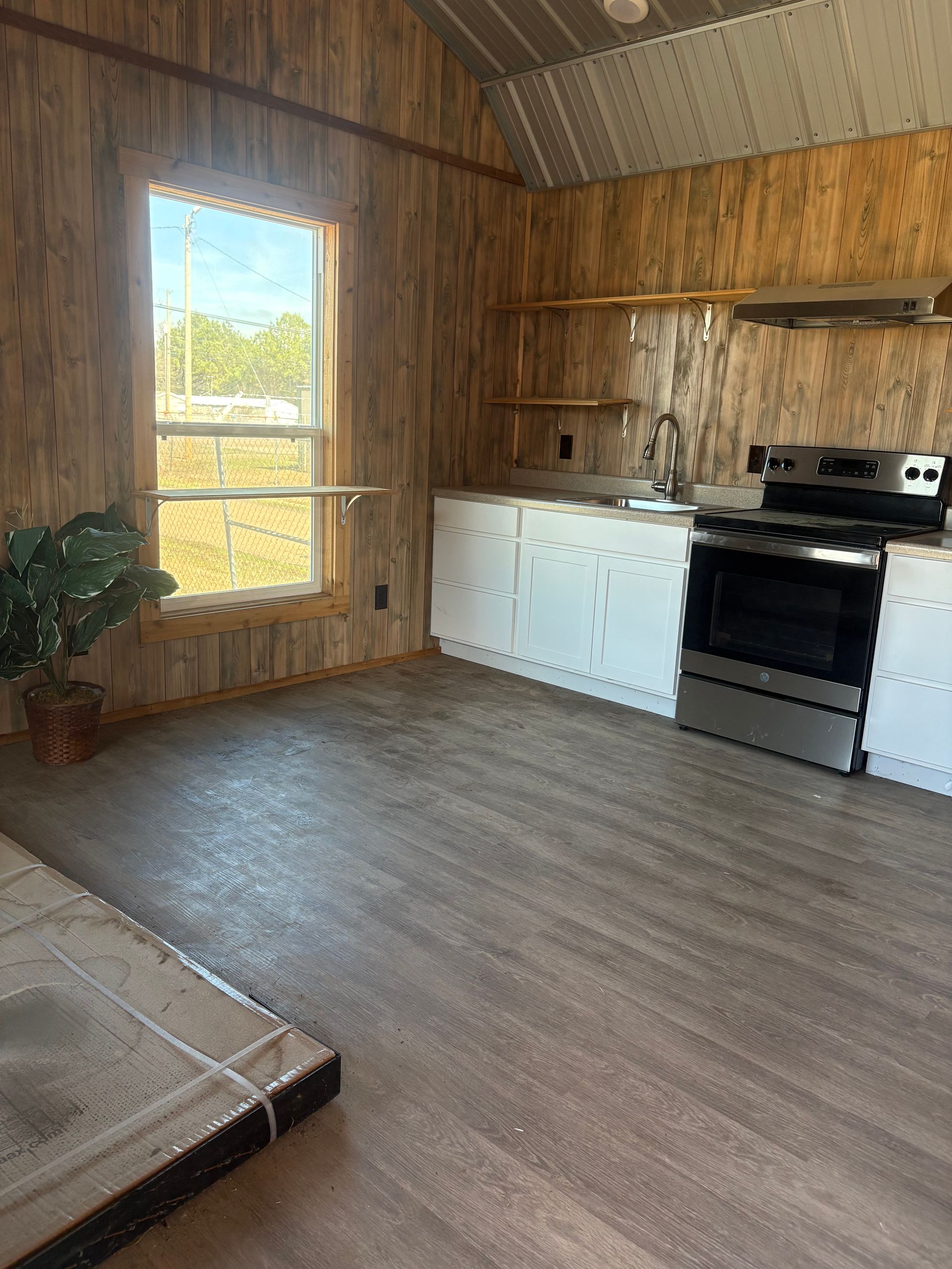 Interior view of a small kitchen. Features white cabinets, a stove, a window, and wood panel walls.