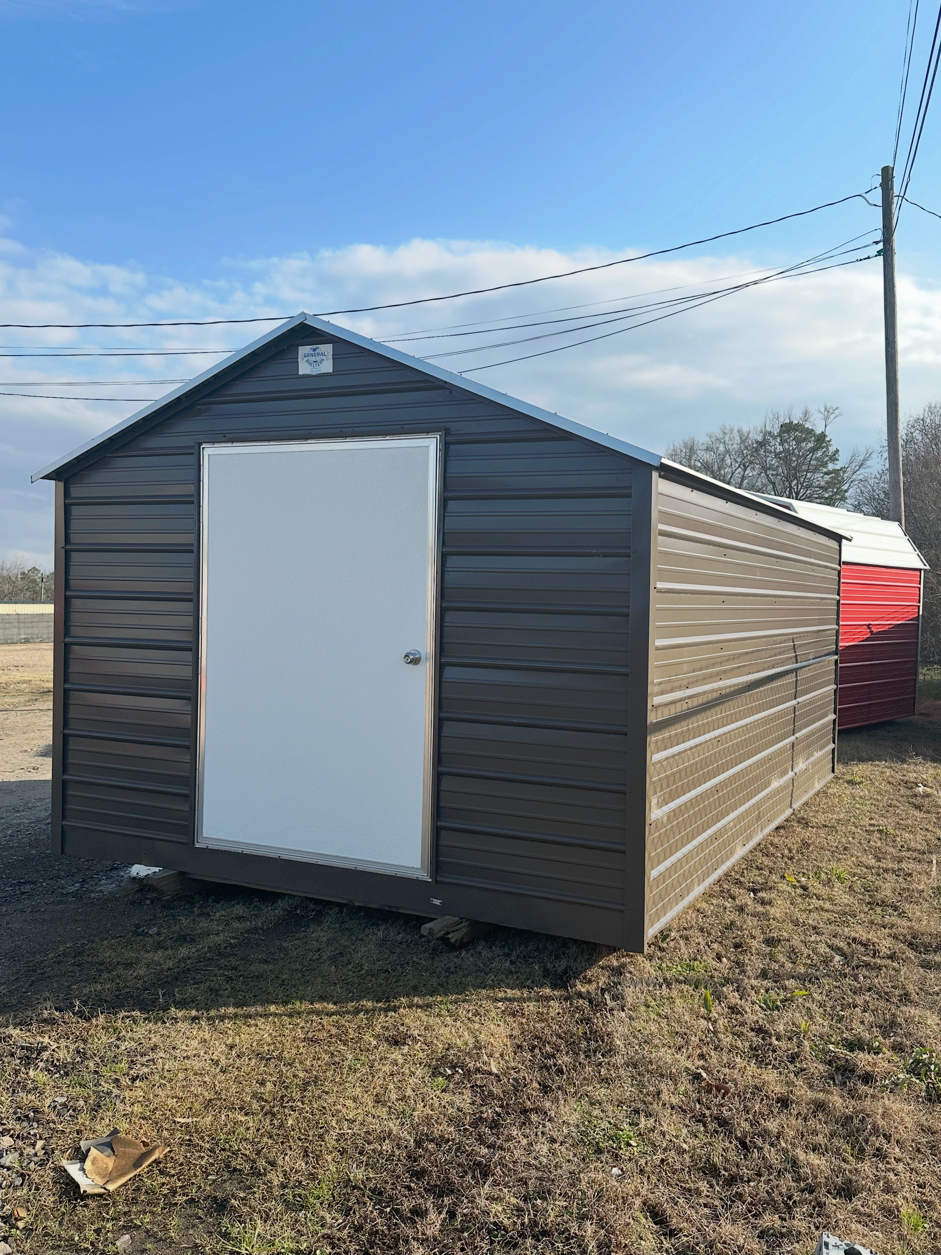 Brown shed with white door, set in a field under a blue sky, with utility poles in background.