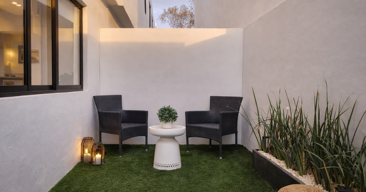 Pequeño patio exterior con dos sillas negras, una mesa pequeña, césped artificial y paredes blancas para casas en Tijuana.