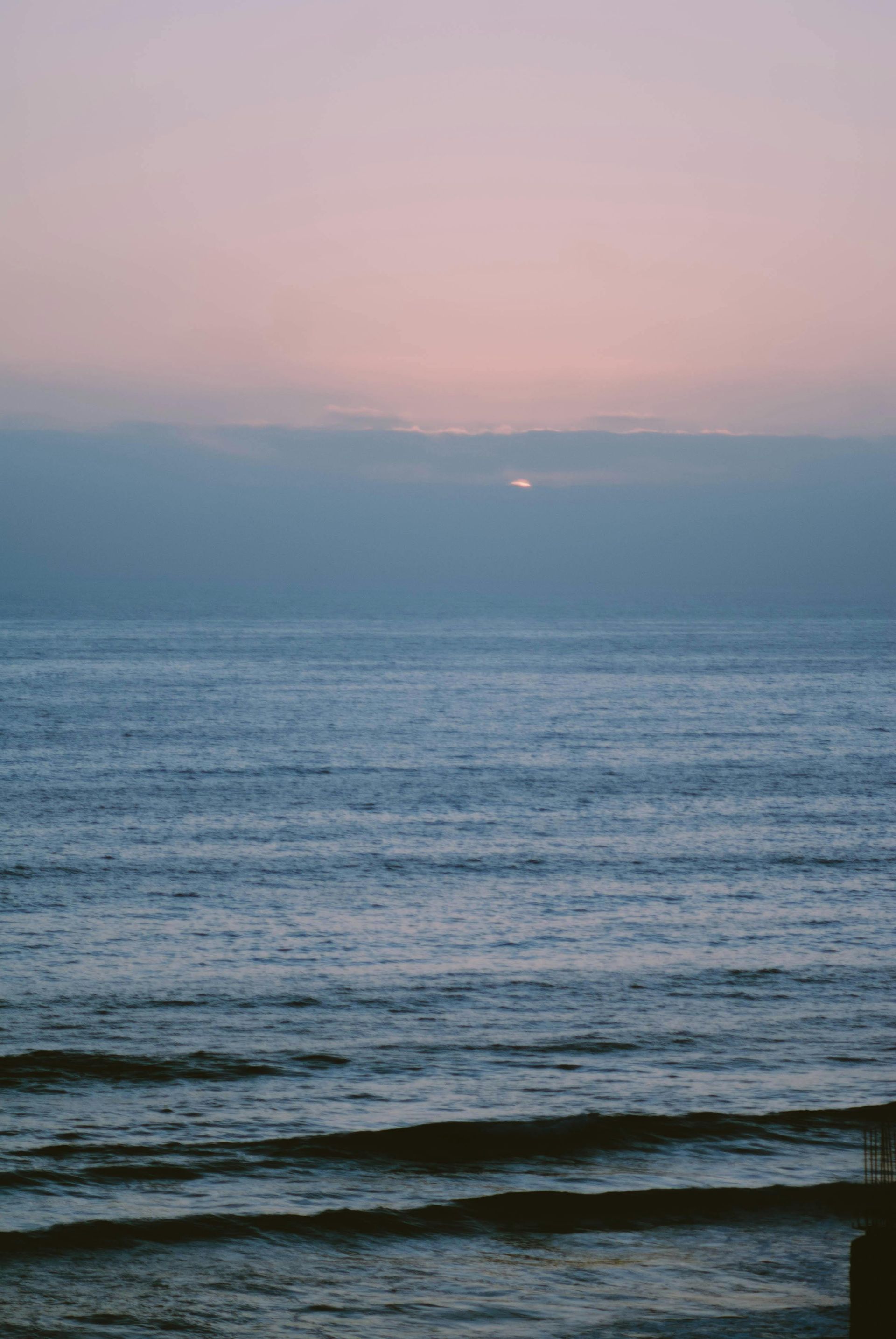 Océano al amanecer, agua azul tranquila bajo un cielo rosa pastel y azul, con un toque de sol asomándose entre las nubes.