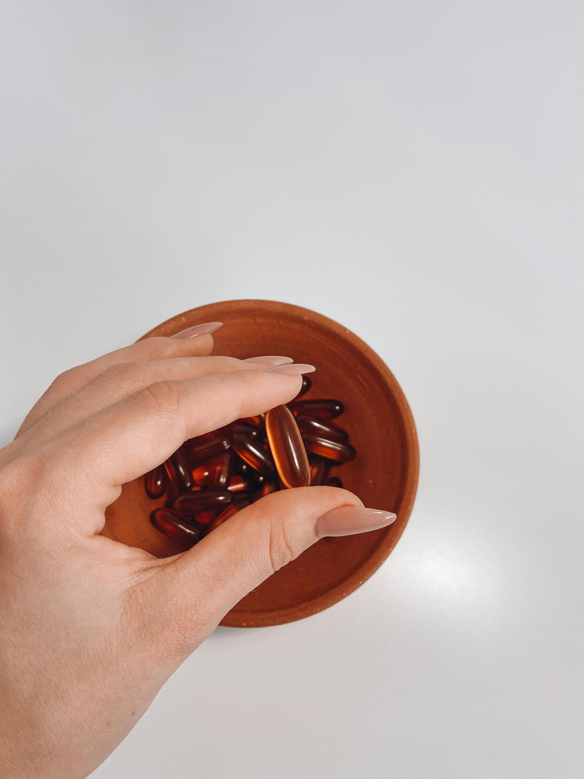 A hand holds a handful of brown capsules in a small wooden bowl on a white surface.
