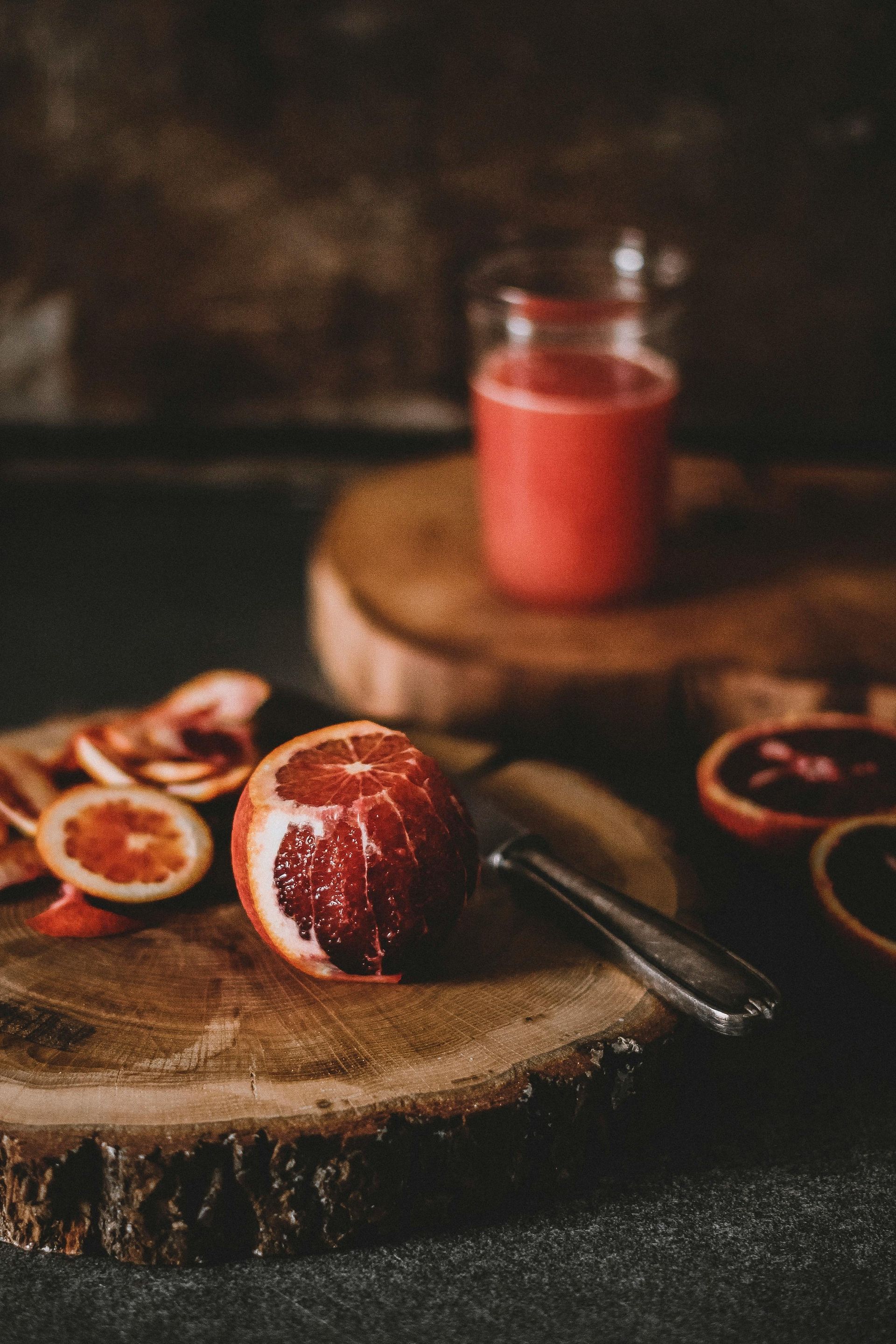 Blood oranges on wooden boards with a glass of juice. Sliced fruit and peeler. Dark, moody lighting.
