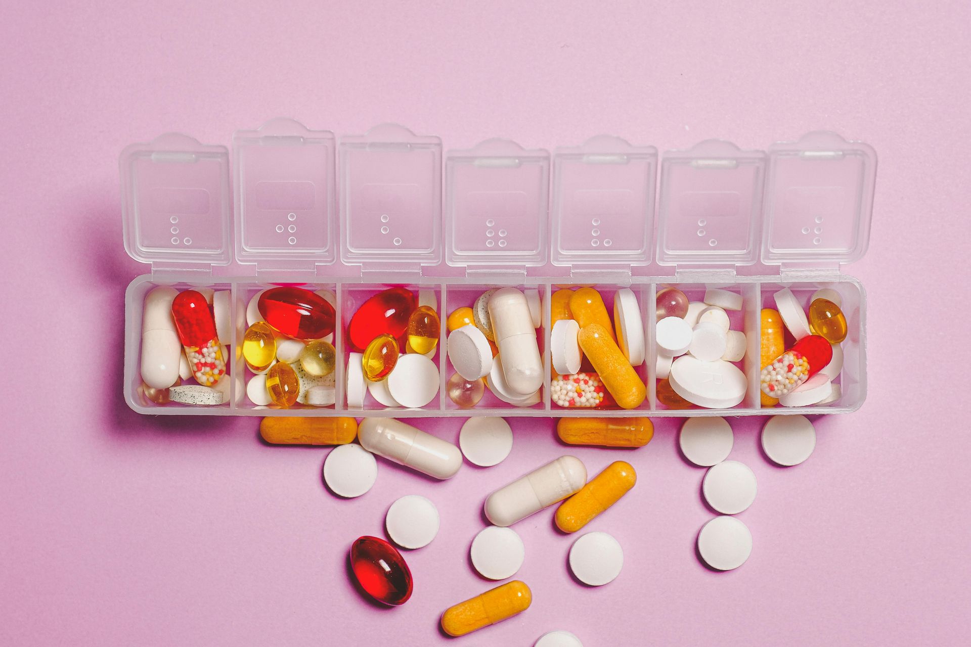A clear plastic pill organizer filled with various pills and capsules on a pink surface. Some pills are scattered outside the organizer.