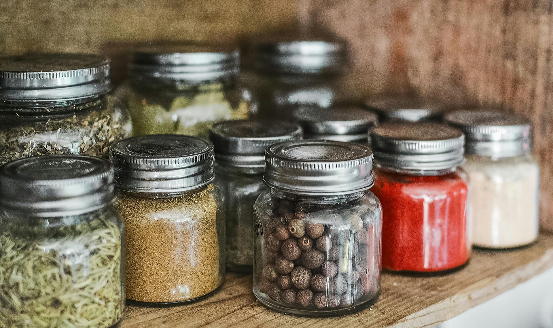 Glass jars filled with spices on a wooden shelf, including green herbs, red powder, and peppercorns.