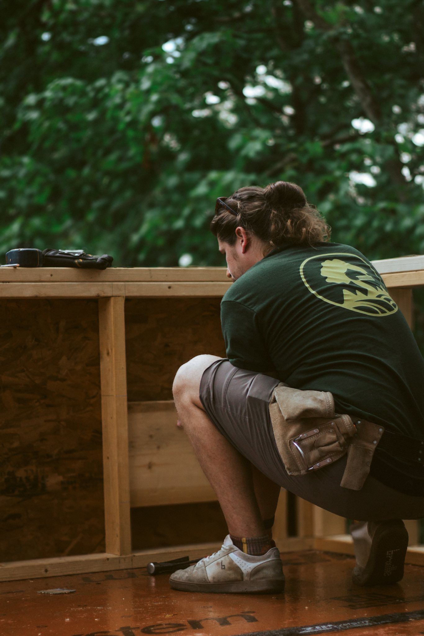 Construction worker squats, working on wooden structure. Dark green shirt, shorts, cloudy day.