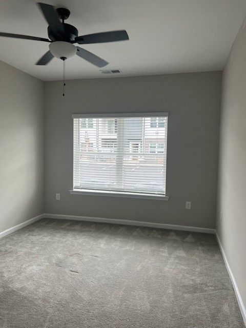 Empty bedroom with gray carpet, walls, and blinds. Ceiling fan and window.