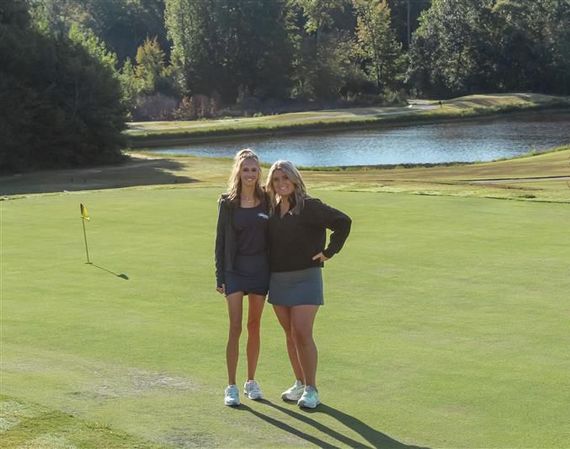 Two women standing on a golf green with a pond and trees in the background.
