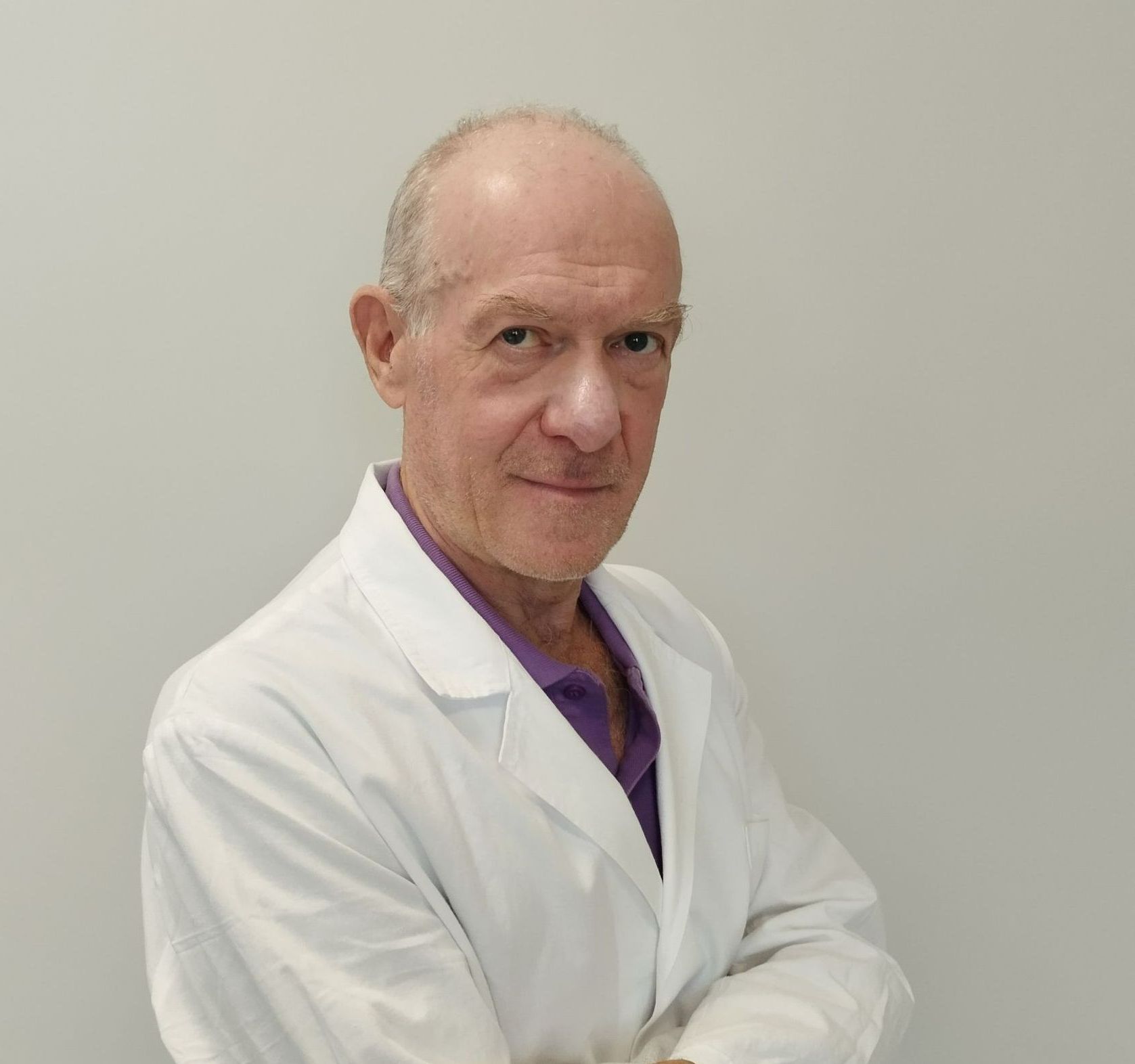 Un uomo con un camice da laboratorio bianco e una maglietta viola è in piedi con le braccia incrociate.