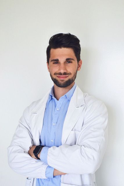 Un uomo con la barba indossa un camice bianco e una maglietta blu.