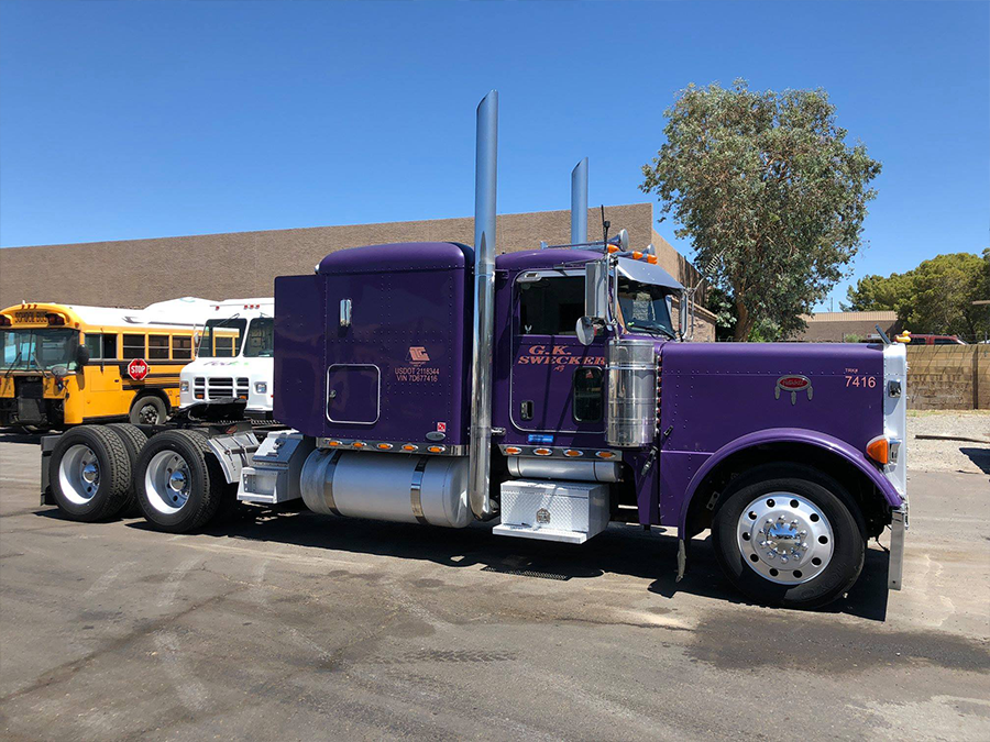 A purple semi truck is parked in front of a row of school buses.