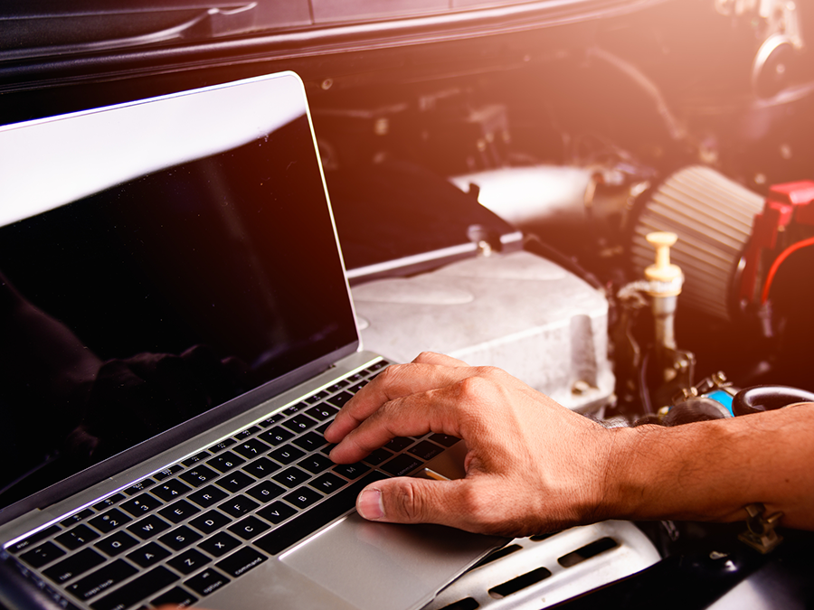 A person is typing on a laptop computer in front of a car engine.