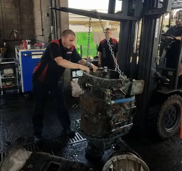 Mechanic using a forklift to move a large engine in a repair shop. Another mechanic observes.