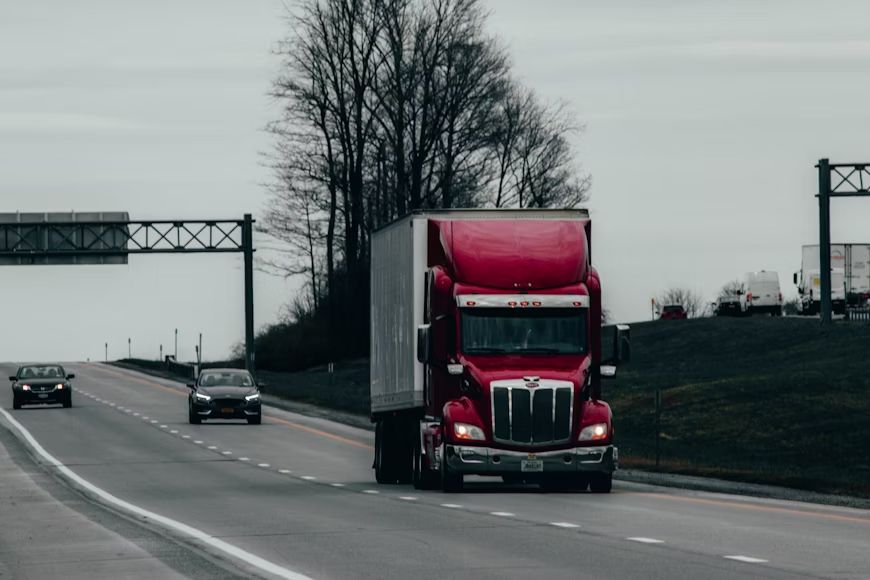 Red semi-truck on a highway, driving toward the viewer under a cloudy sky.