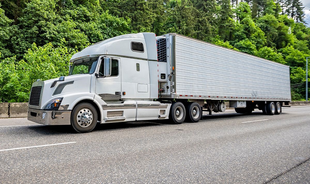 White semi-truck on a gray road next to green trees and foliage.