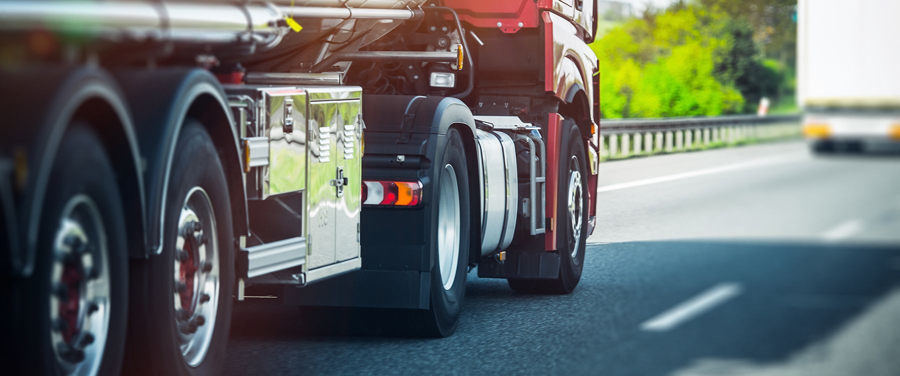 Red semi-truck driving on a highway beside a guardrail, with another truck in the distance.