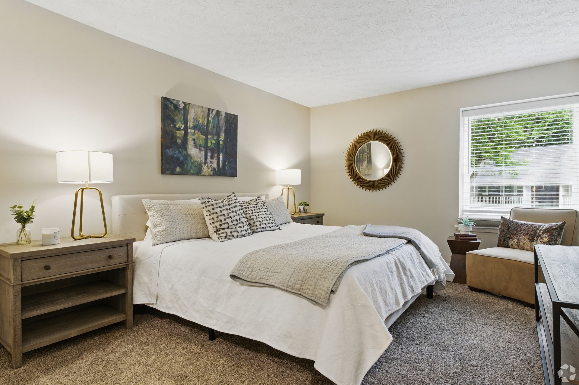 Bedroom with bed, nightstands, artwork, mirror, chair, and window. Neutral colors, soft lighting.