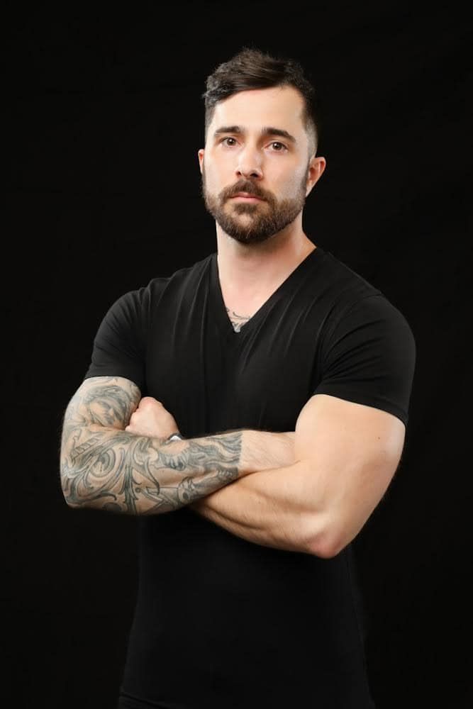 A man with a full beard and tattooed arm, wearing a black v-neck shirt, stands with arms crossed against a black background.