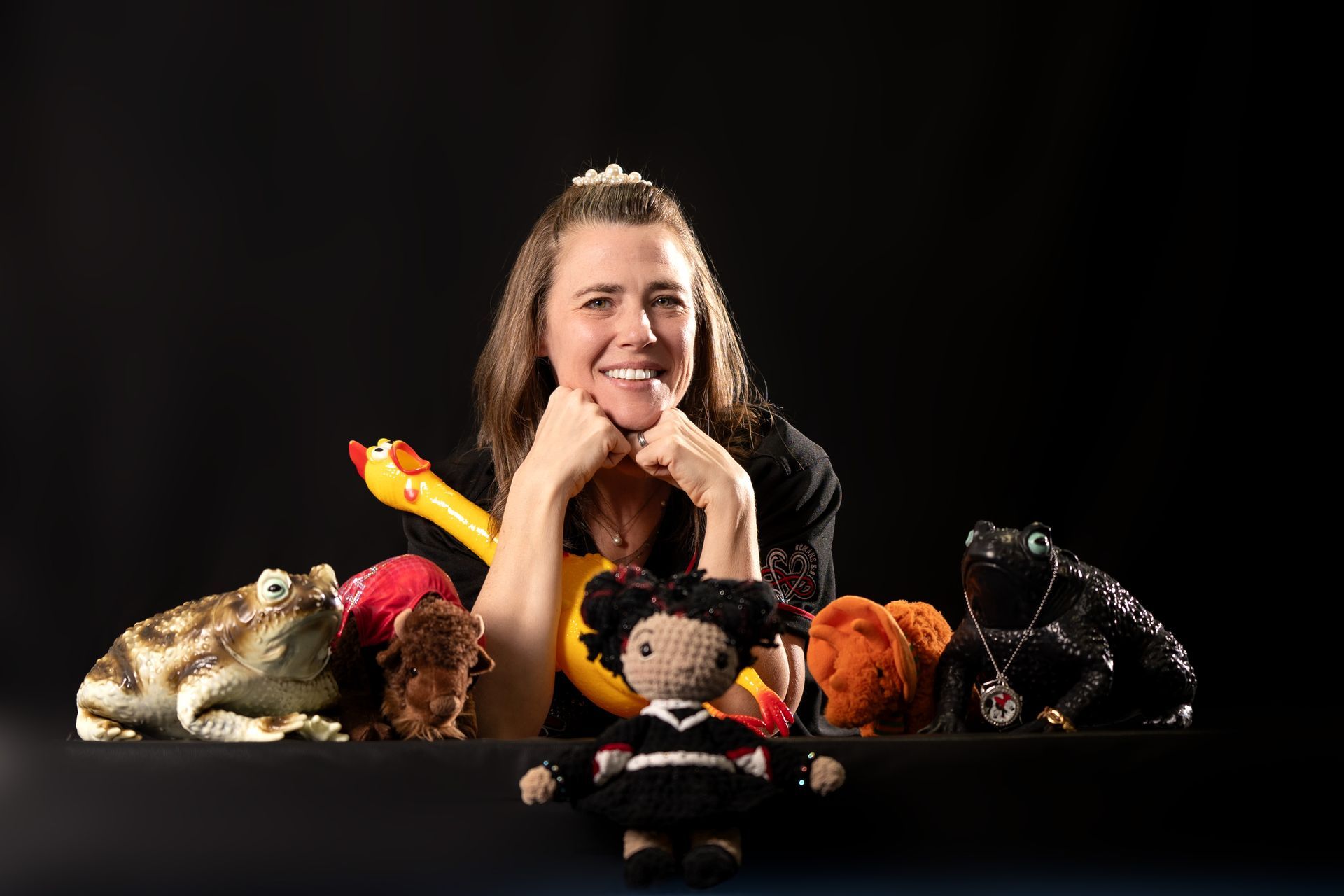 A person posing behind a black table with a rubber chicken, a crochet doll, two frog figurines, and other plush toys.