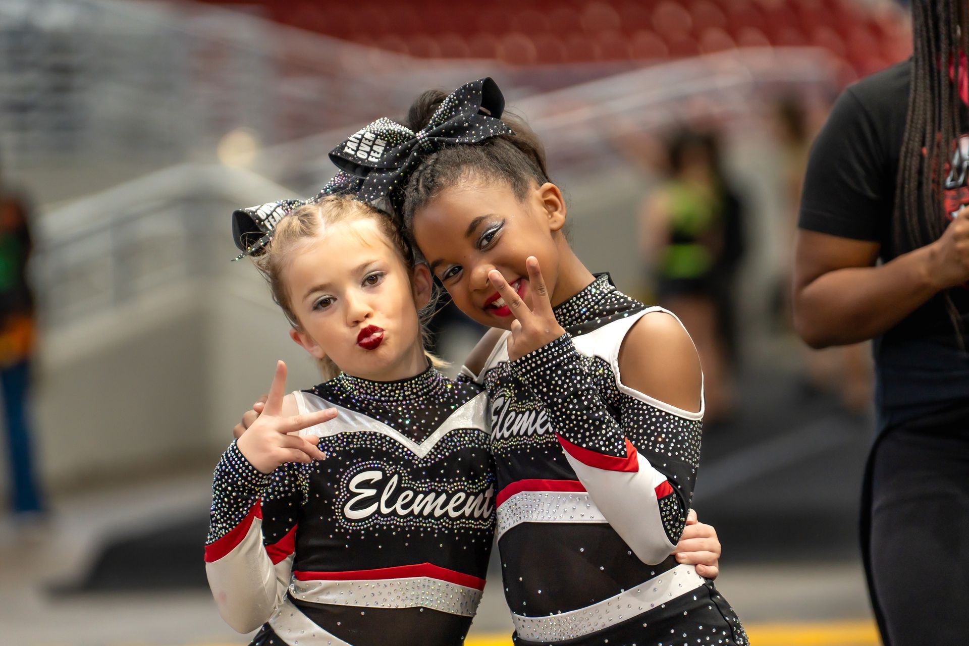 Two cheerleaders in matching black and white uniforms with large bows posing and making hand signs in an arena.