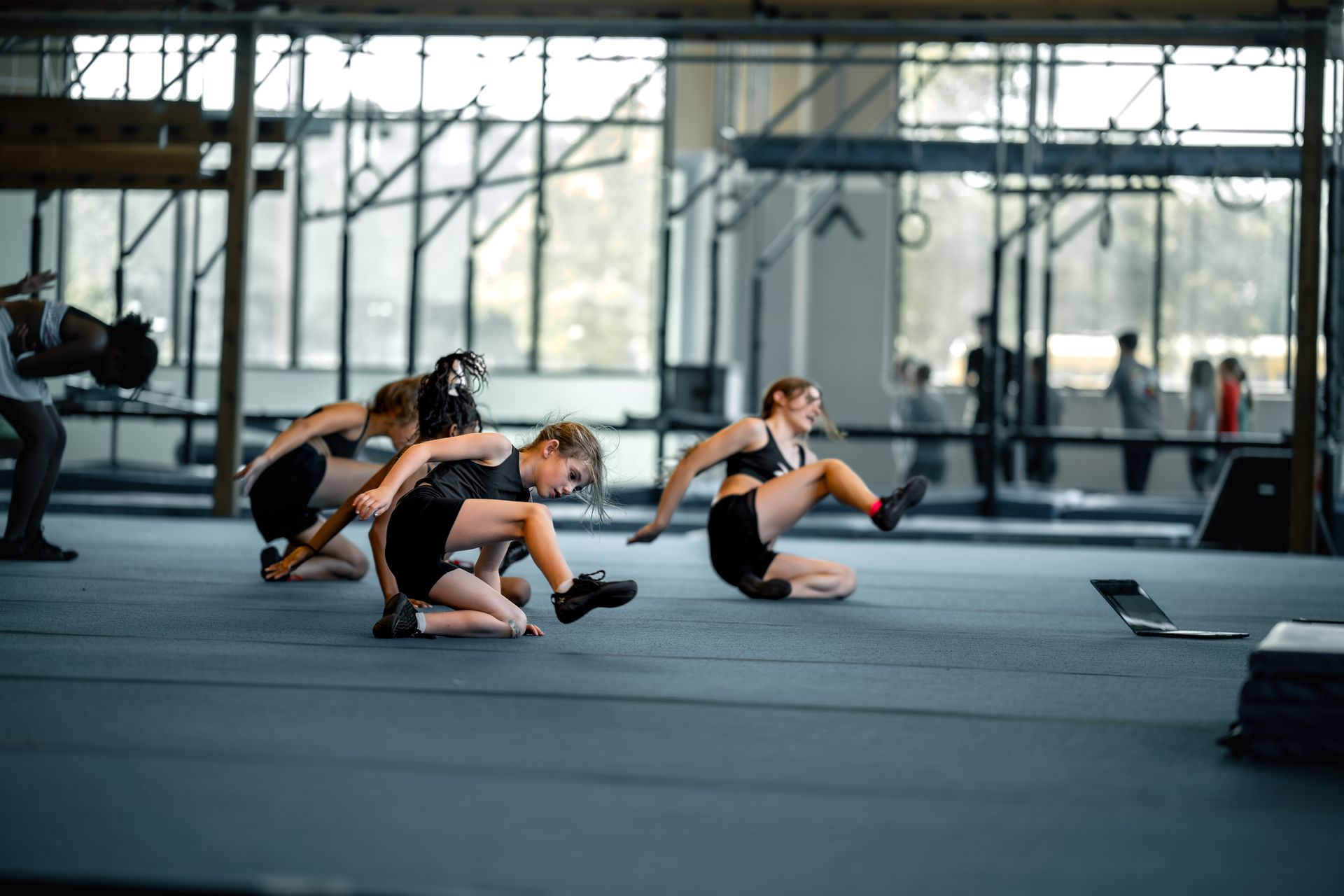 Three dancers in black athletic wear perform a floor routine in a large, mirrored gym with industrial ceilings.