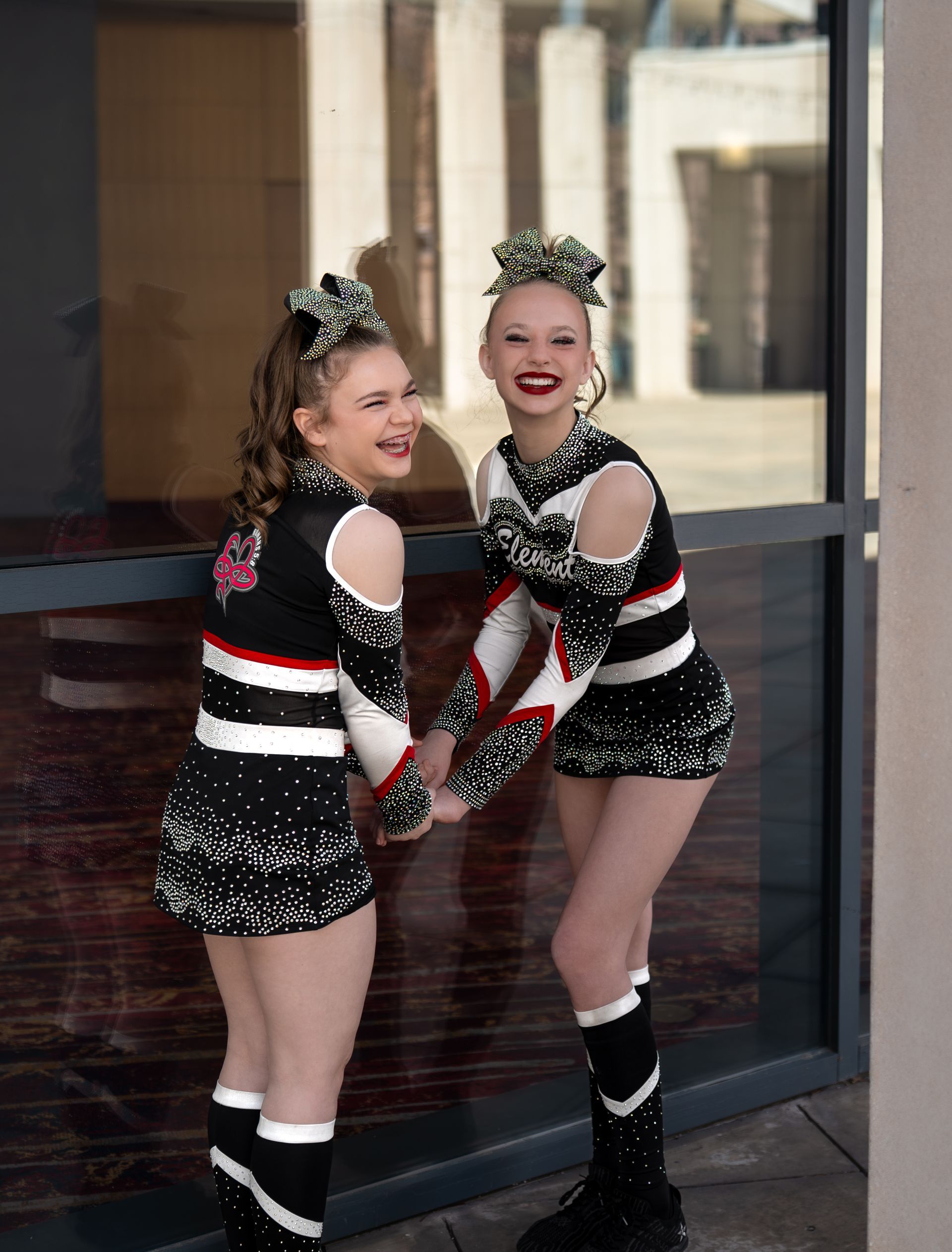 Two people in matching black, white, and red cheerleading uniforms and hair bows, laughing while holding hands outdoors.