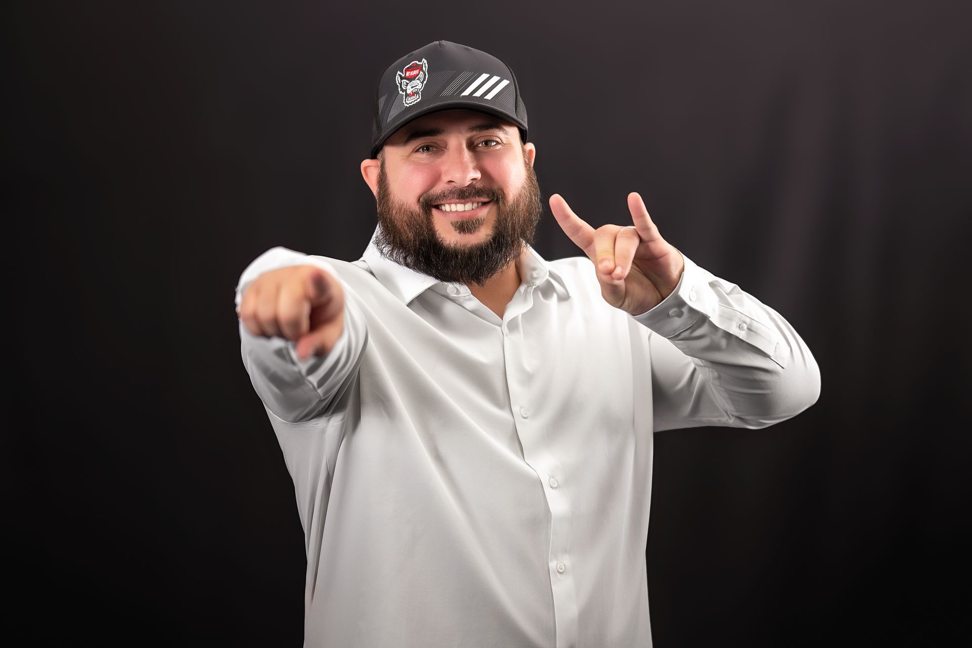 A smiling person in a white button-down shirt and a black baseball cap pointing forward with one hand and gesturing.