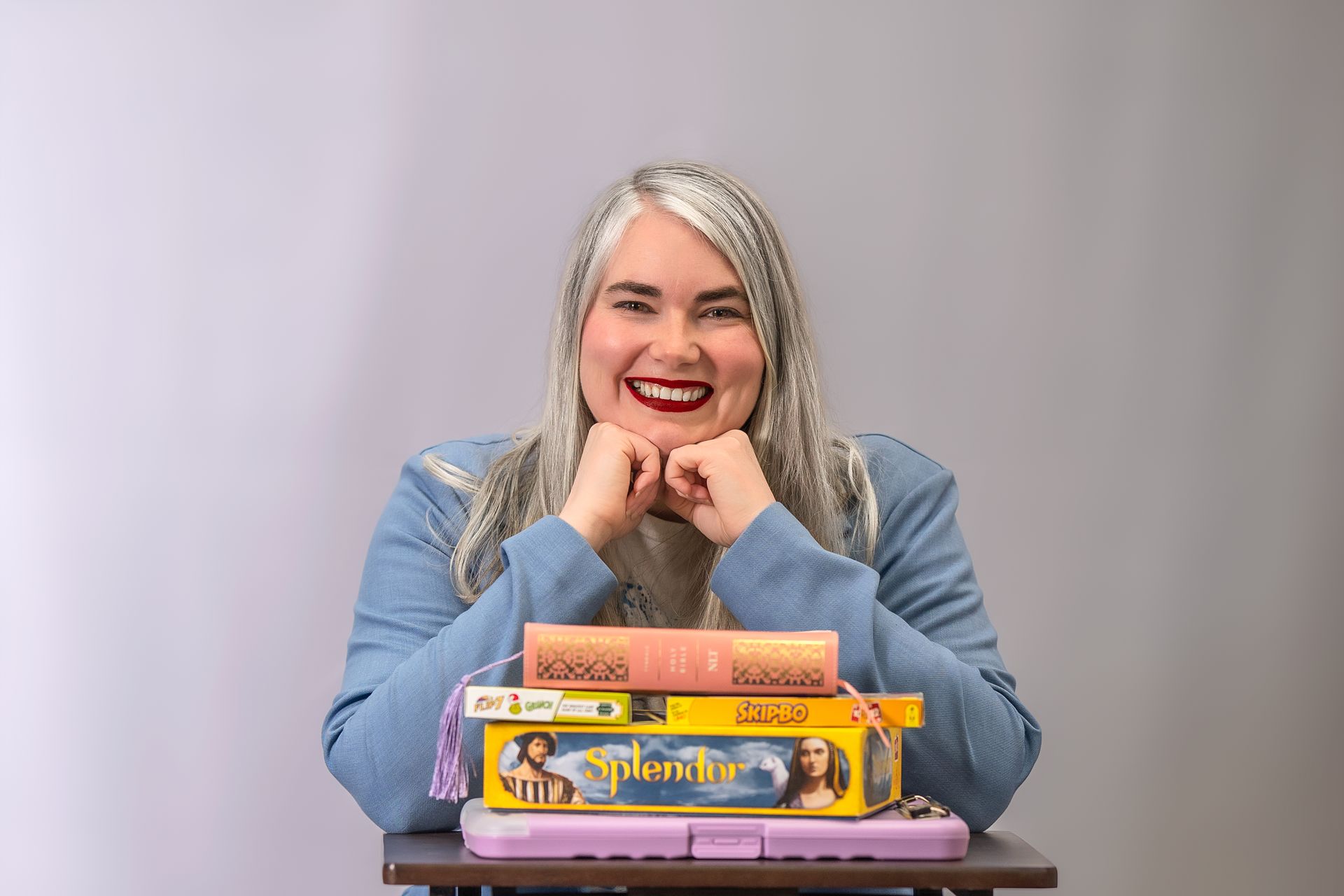 A person smiles with their chin resting on their hands behind a stack of board games on a desk against a grey background.