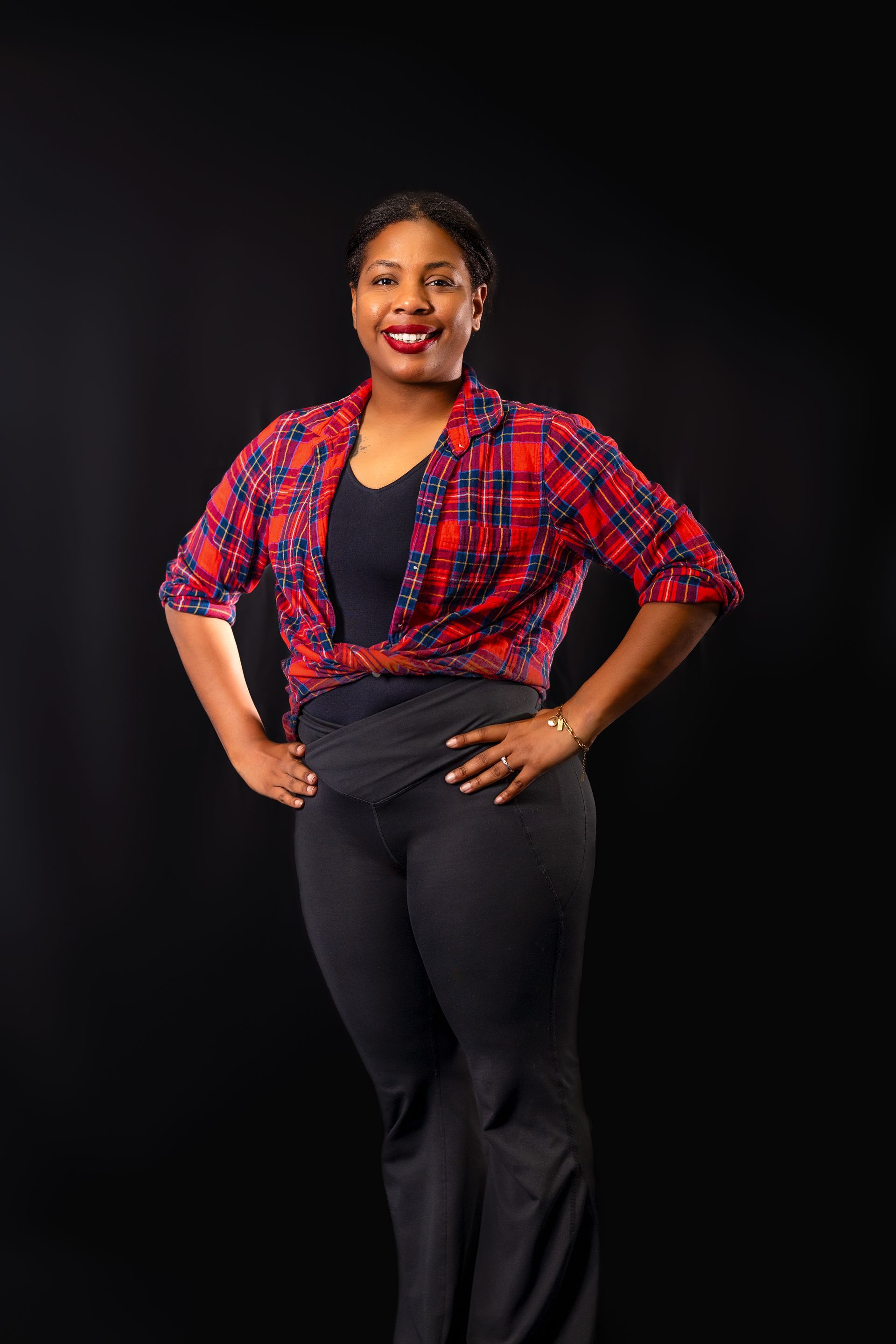 A person posing with hands on hips, wearing a red plaid shirt over a black top and dark jeans against a black background.