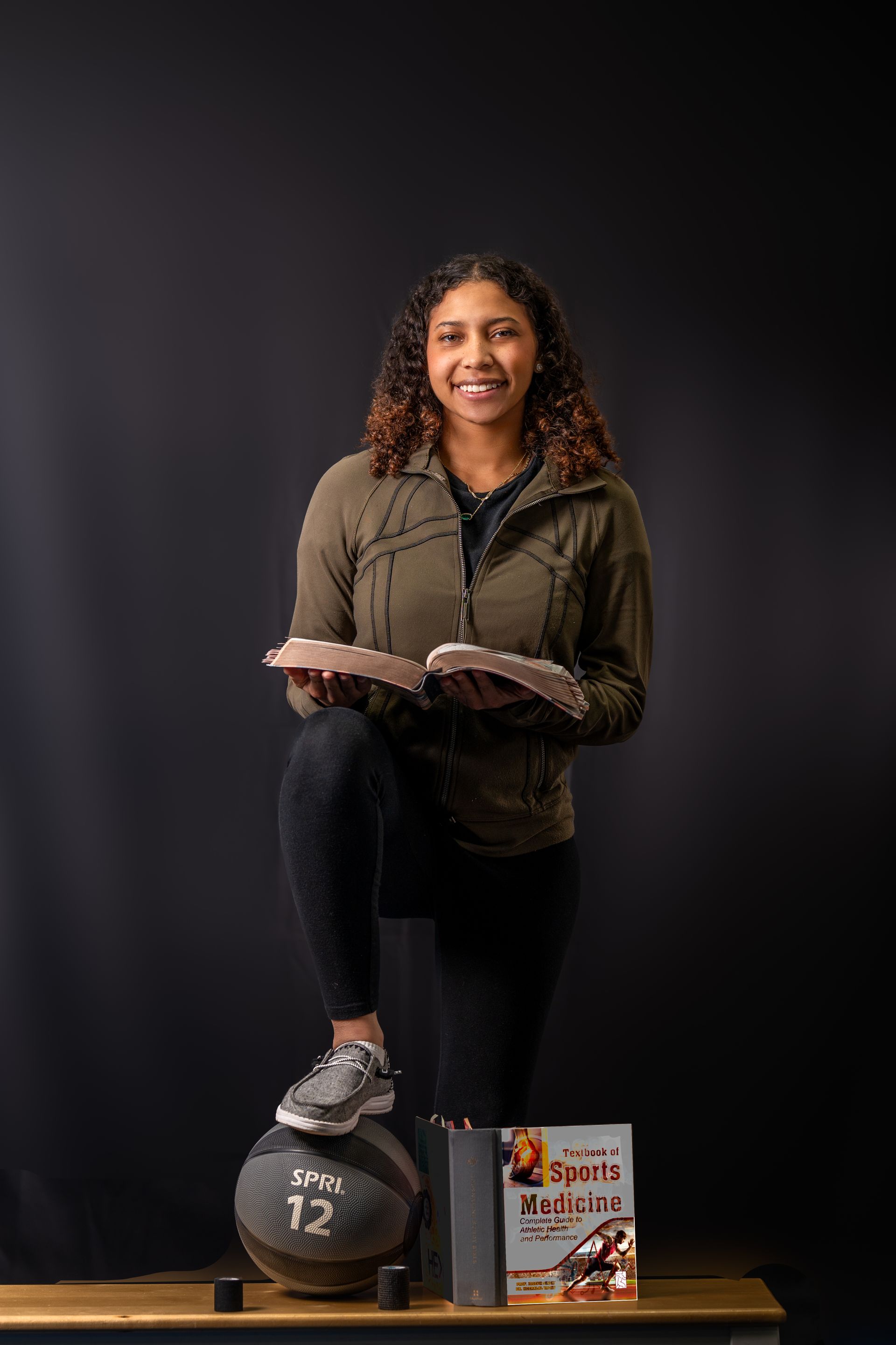 A person smiles while holding a book, resting one foot on a number 12 medicine ball beside a book display on a table.