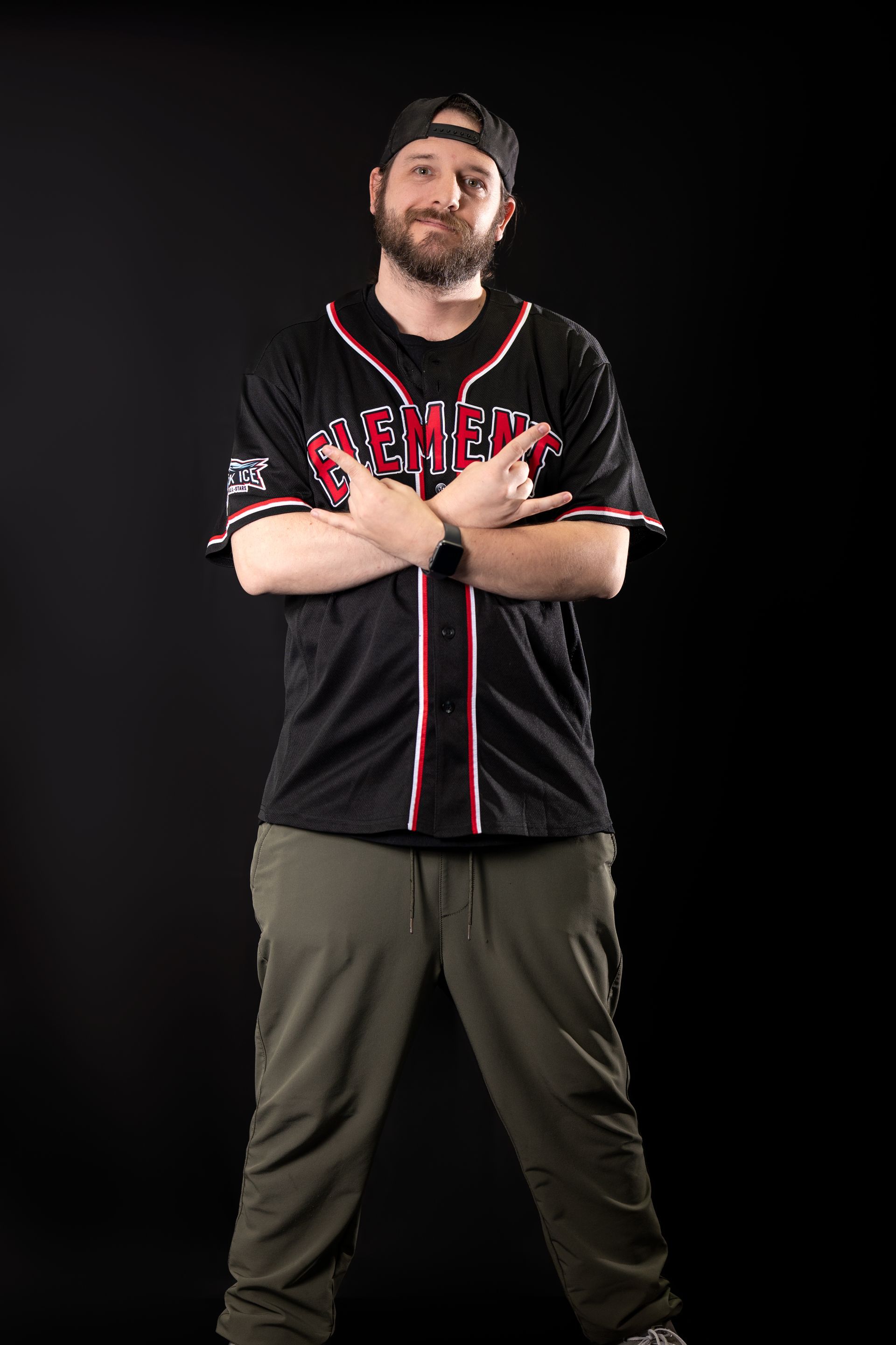 A bearded individual in a black baseball jersey and green pants stands against a black background, making a hand gesture.