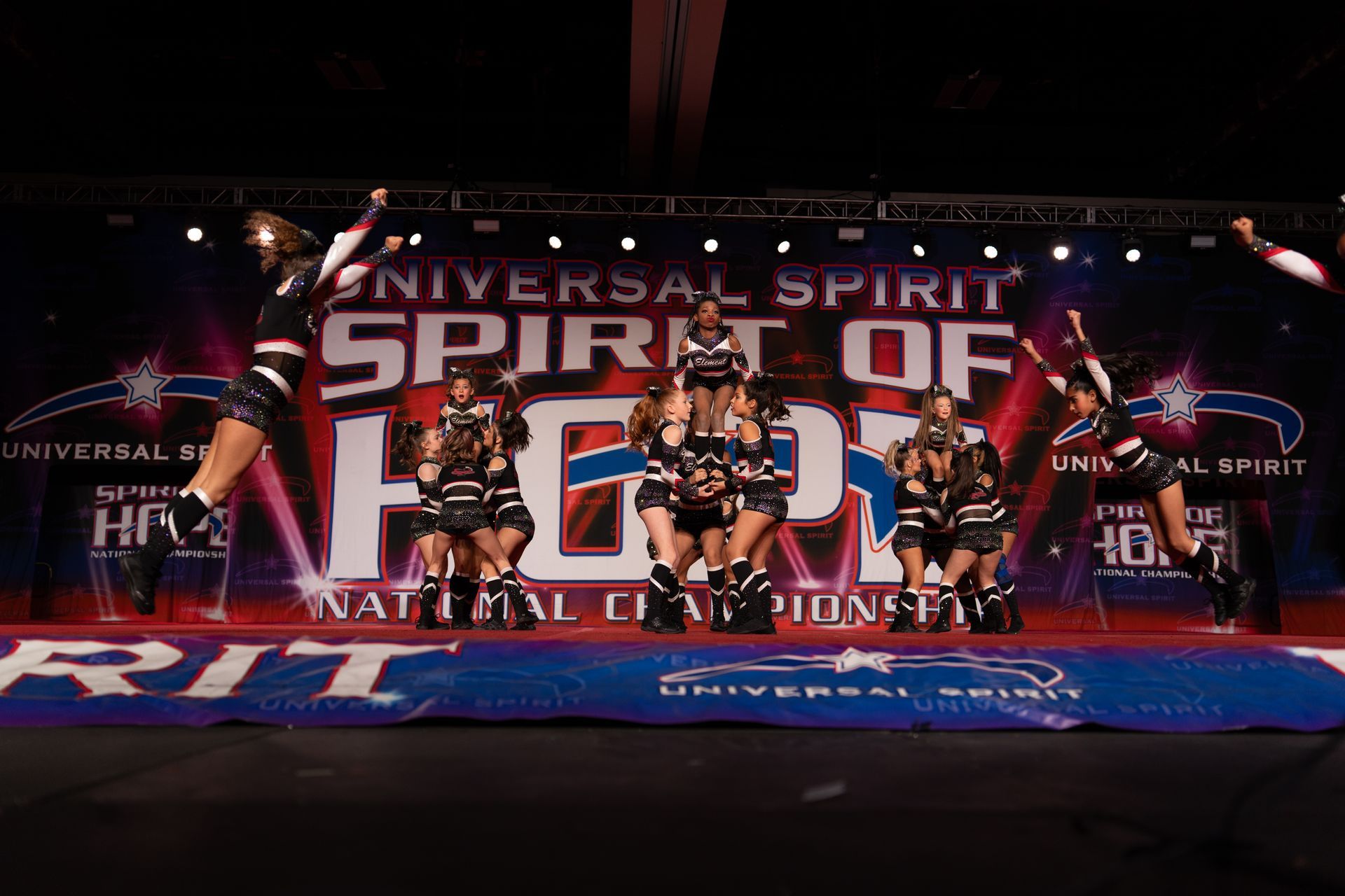 A cheerleading squad performs in coordinated outfits on a stage in front of a 