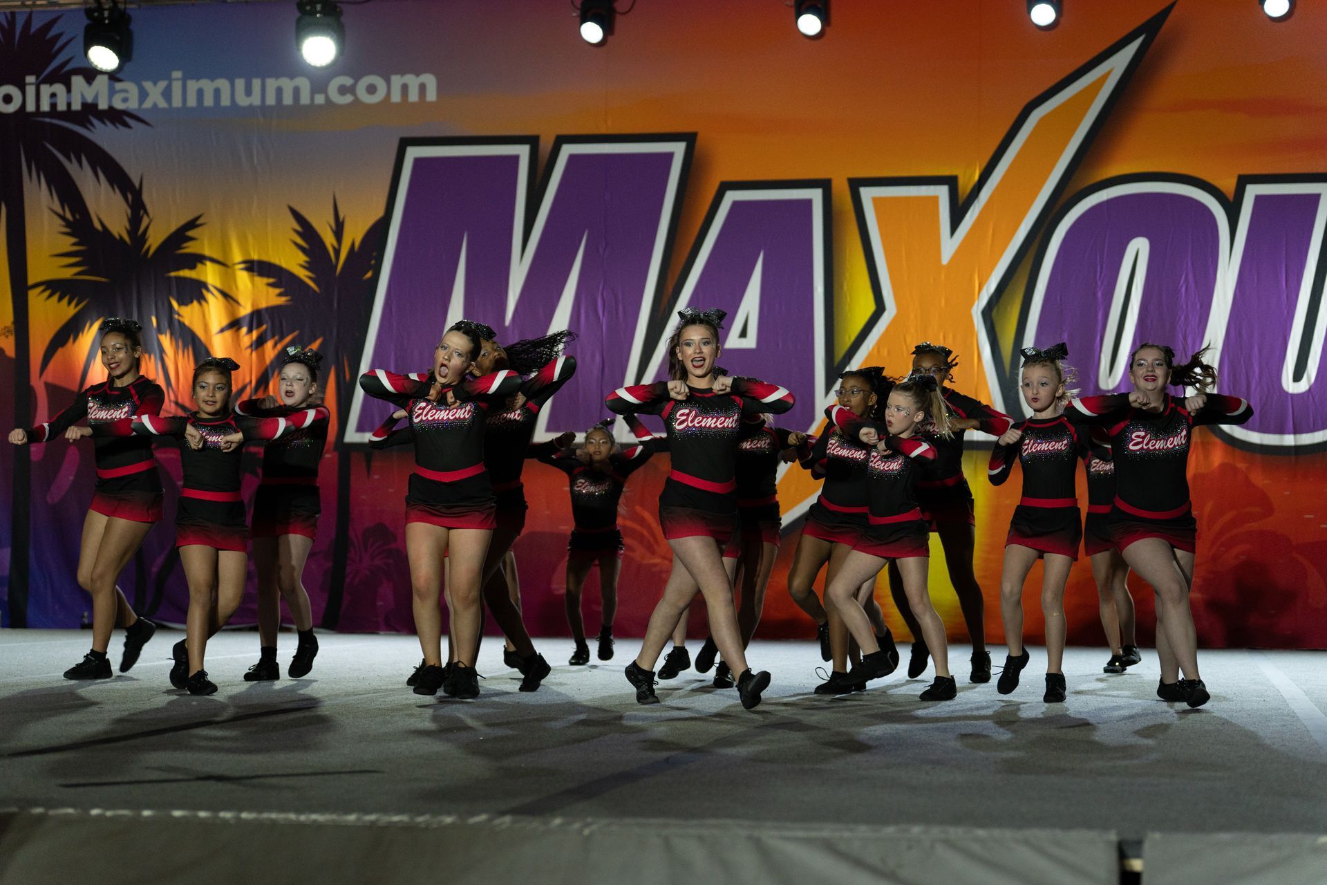 A group of cheerleaders in black and red uniforms performing a choreographed dance on a stage with a sunset backdrop.