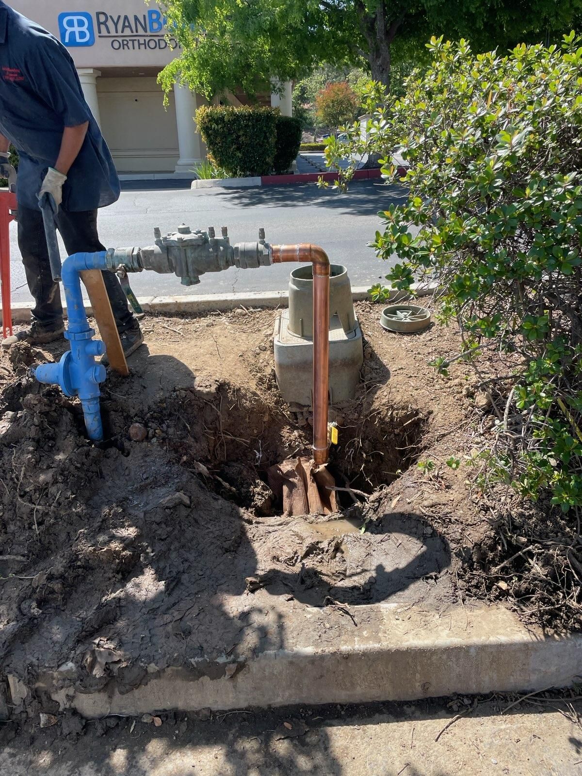 Plumber working on water meter.  Blue and copper pipes in an open dirt hole near a bush and curb.