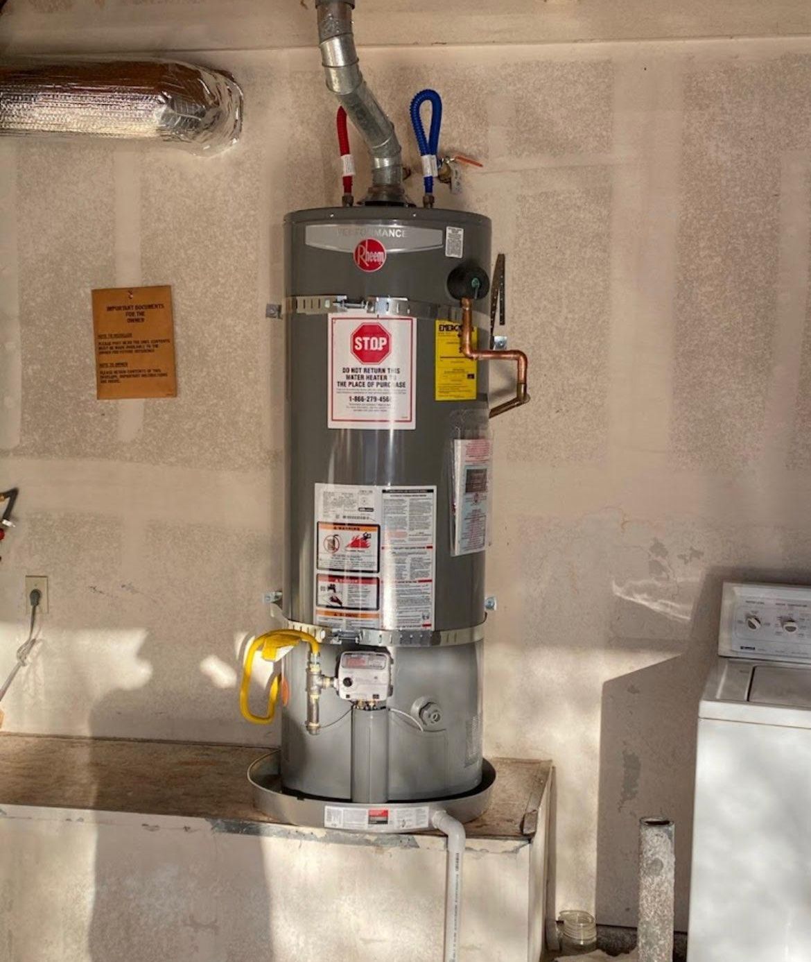 A Rheem water heater in a garage, with water pipes and a yellow gas line, next to a washing machine.