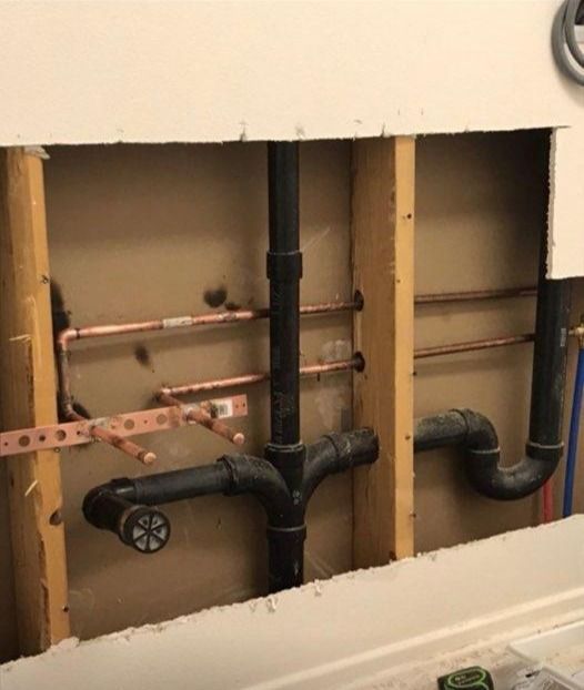 Plumbing exposed in a wall: copper pipes, black drainpipes, and wooden studs.
