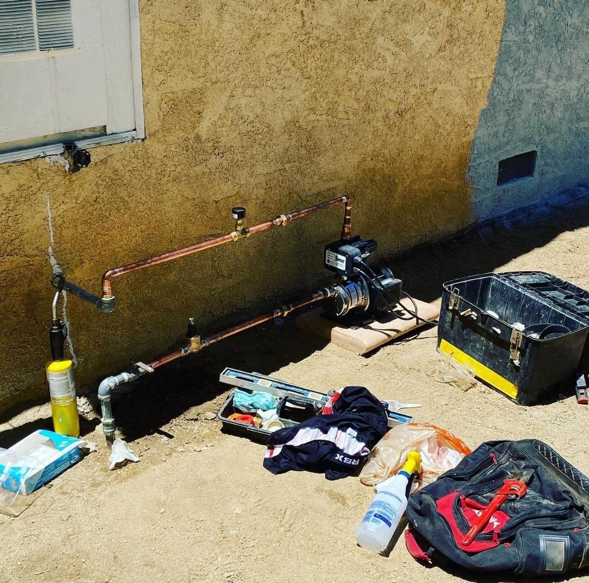 Plumbing work in progress outside. Copper pipes are attached to a water pump. Tools and equipment are scattered around.