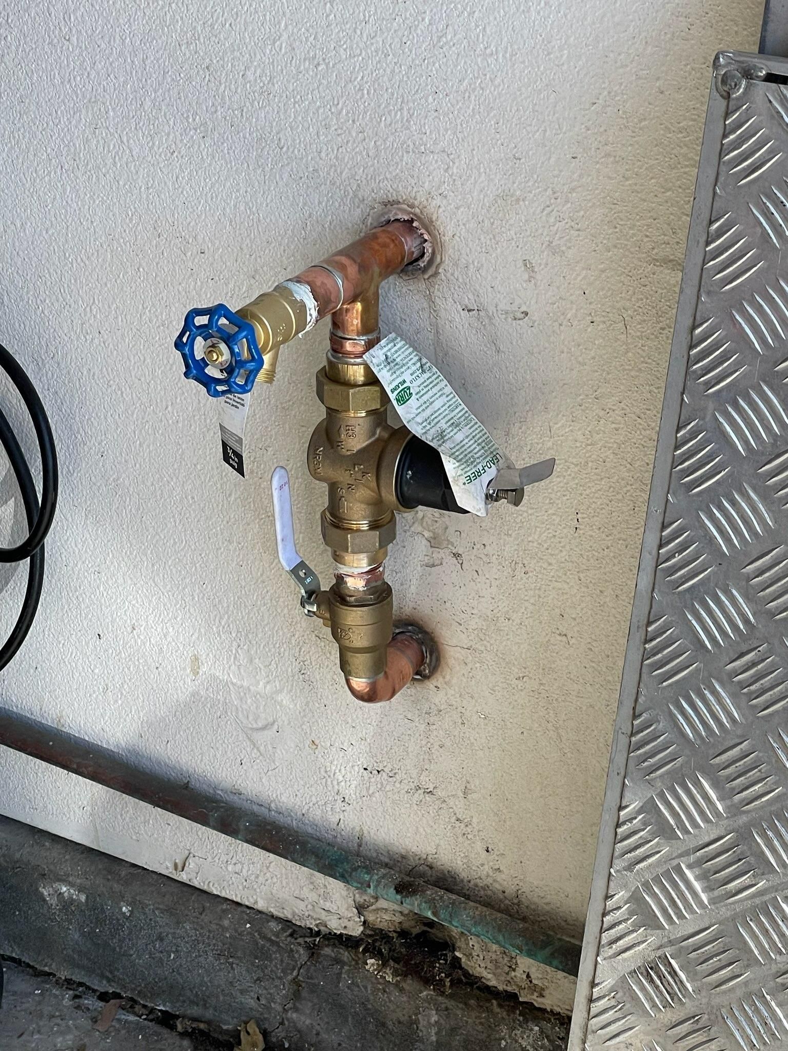 Copper and brass water valve assembly with blue handle, mounted on a concrete surface, next to a metal grate.
