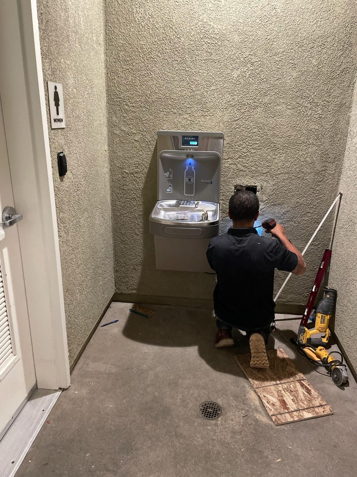 A person installs a water fountain in a restroom. The walls are textured with a gray concrete floor.