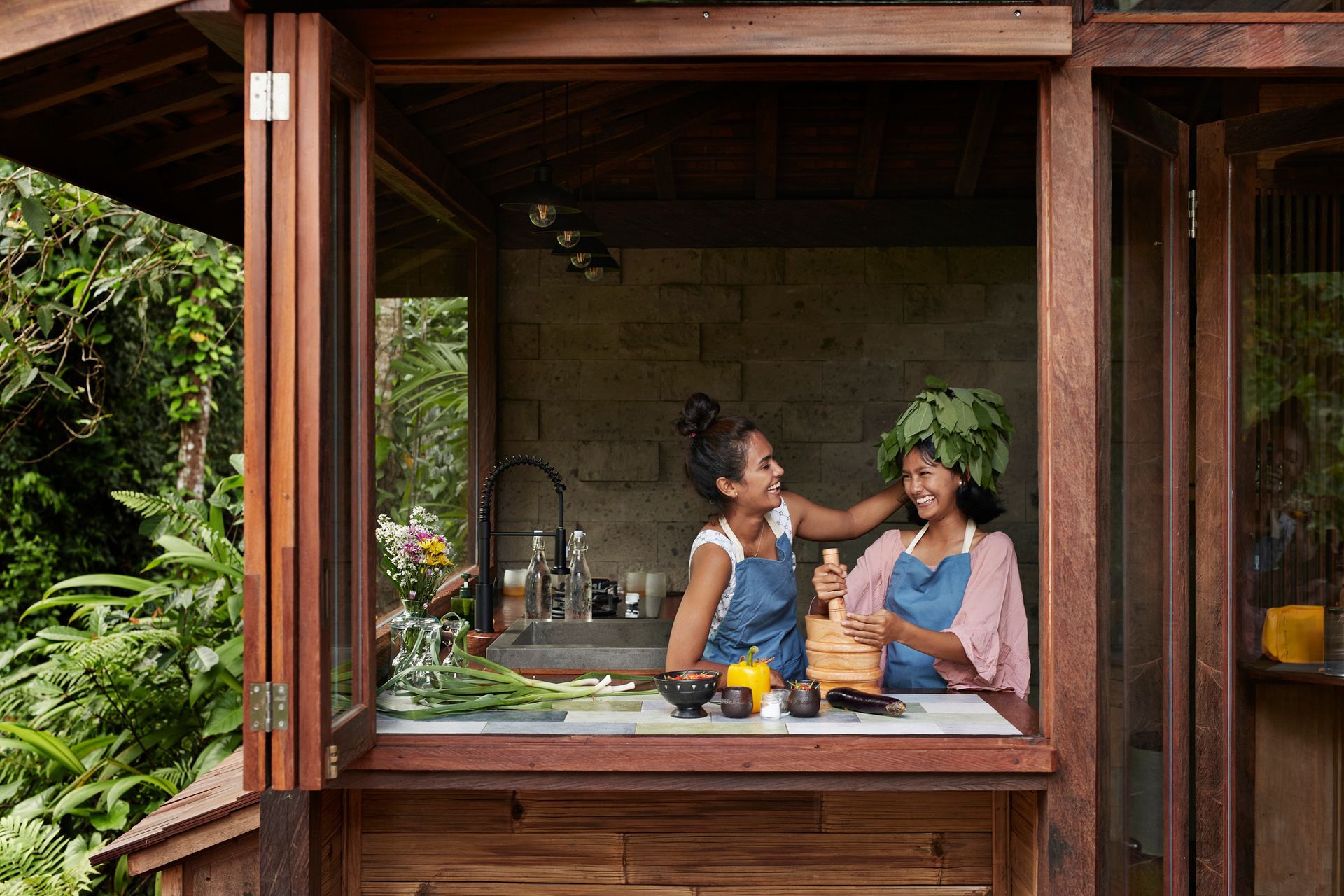 Two women prepare food in a wooden outdoor kitchen with plants, utensils, and a lush green backdrop.