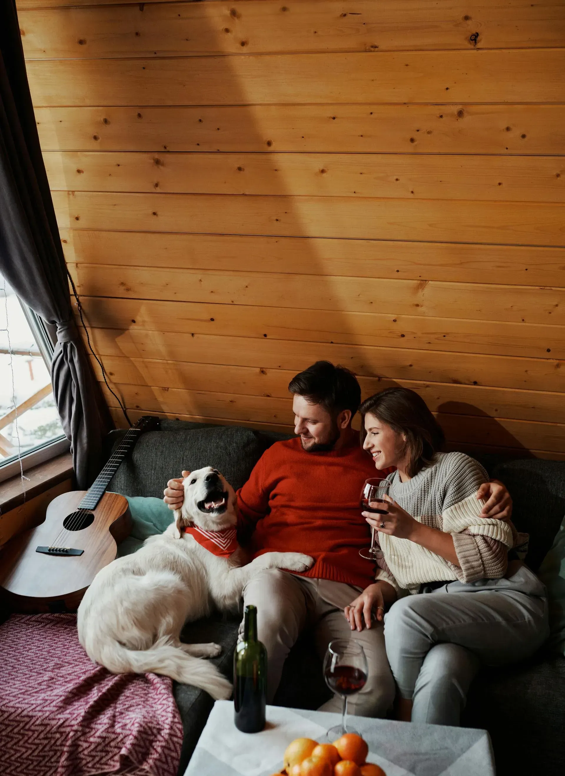 A man and a woman are sitting on a couch with a dog and drinking wine.