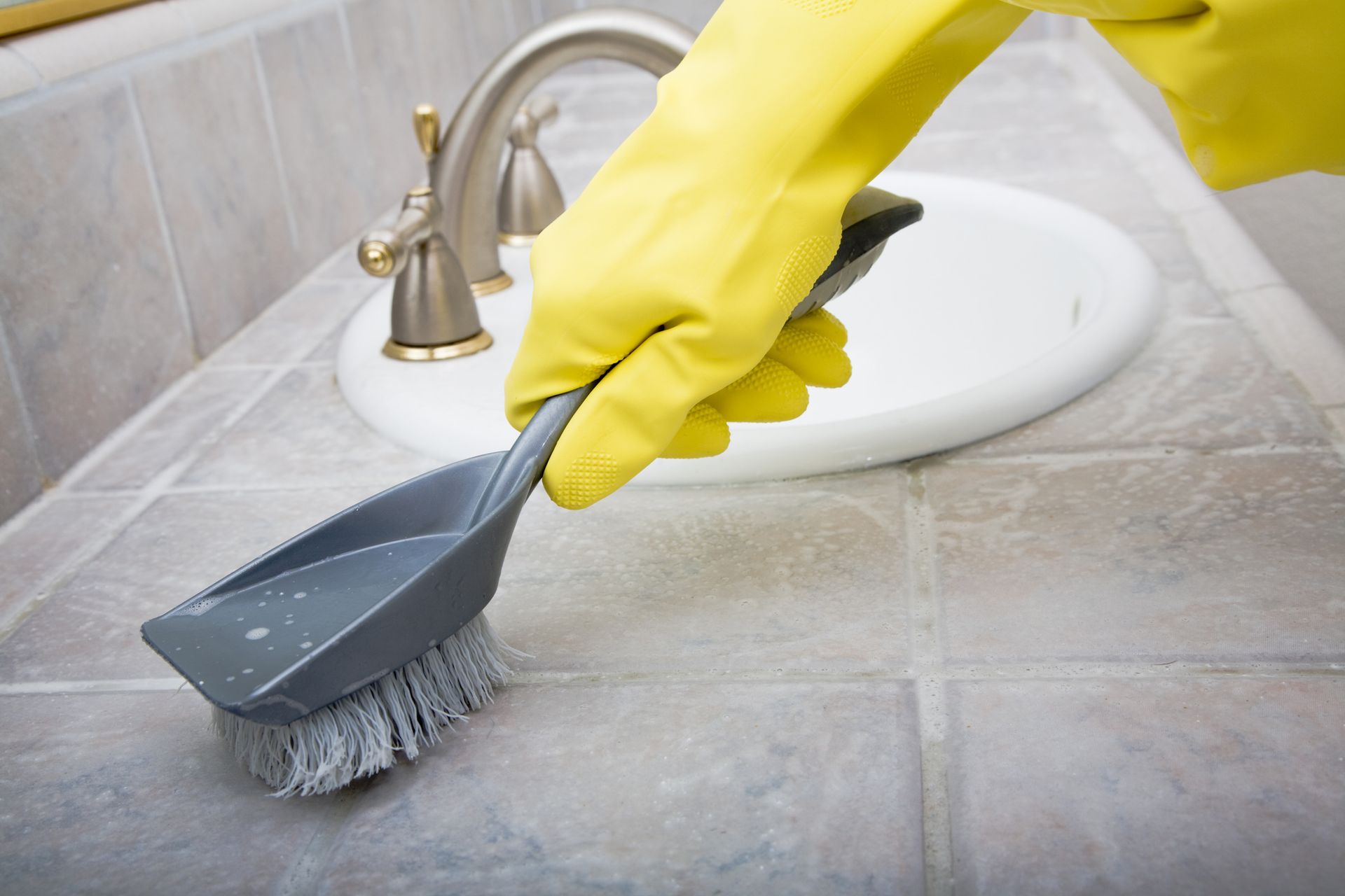 A person wearing yellow gloves is cleaning a bathroom counter with a brush