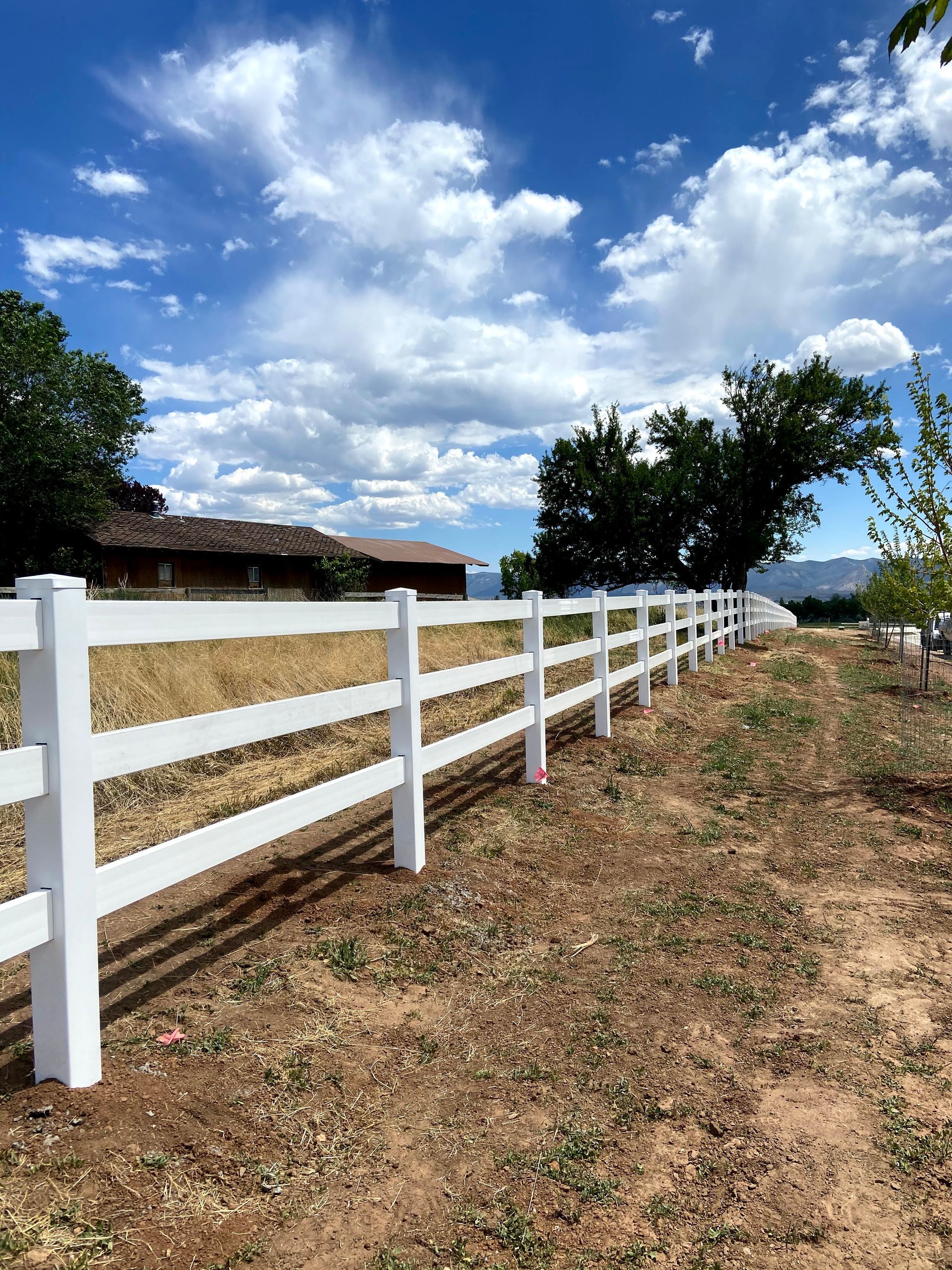 A white fence surrounds a dirt field with a house in the background.